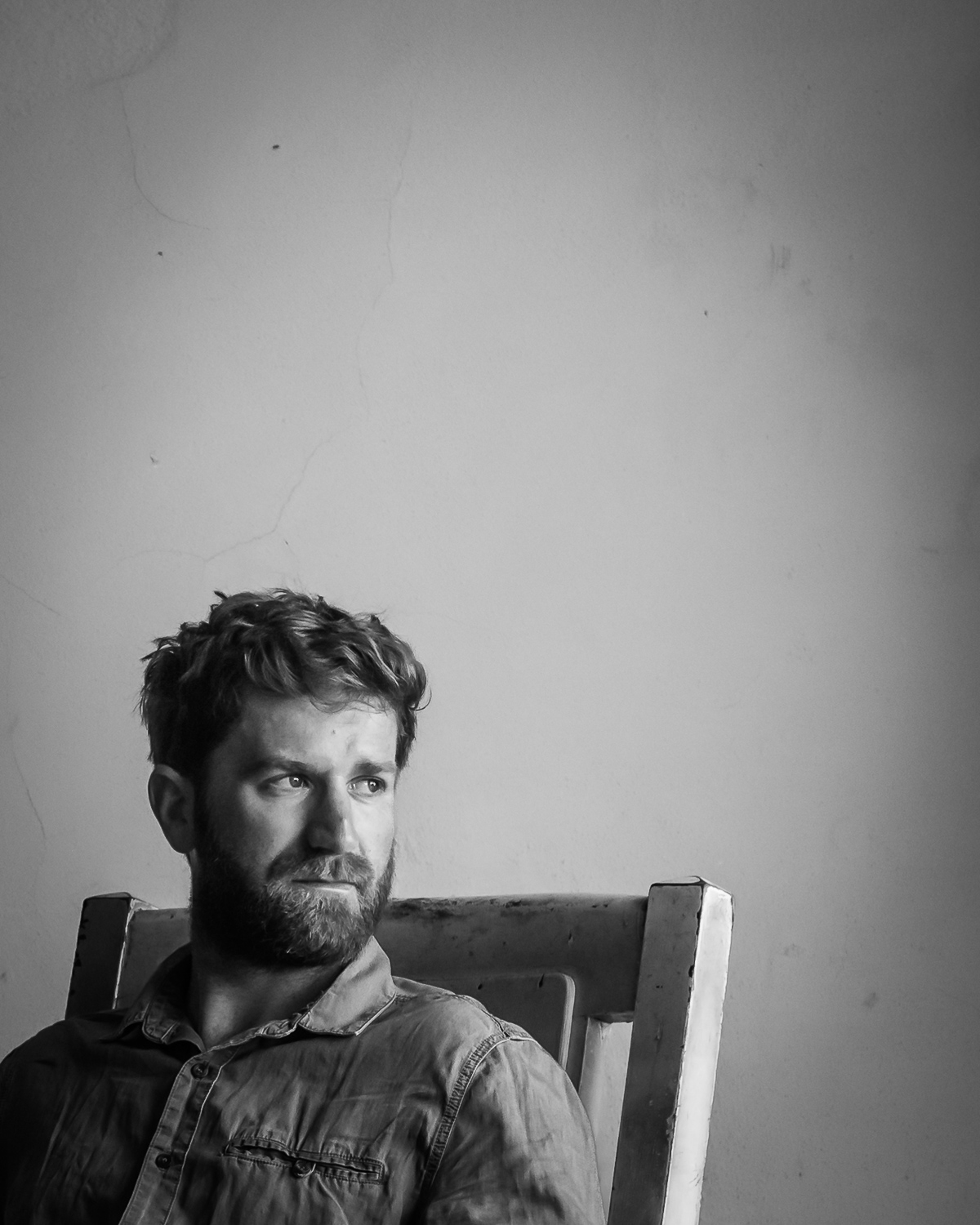 Black and white portrait of Iain, seated in a wooden chair against a bare wall, looking off to one side